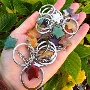 May include: A selection of star-shaped gemstone keyrings in various colours, including green, orange, red, and purple. Each star is attached to a silver keyring with a chain. The keyrings are held in a hand against a backdrop of green leaves.