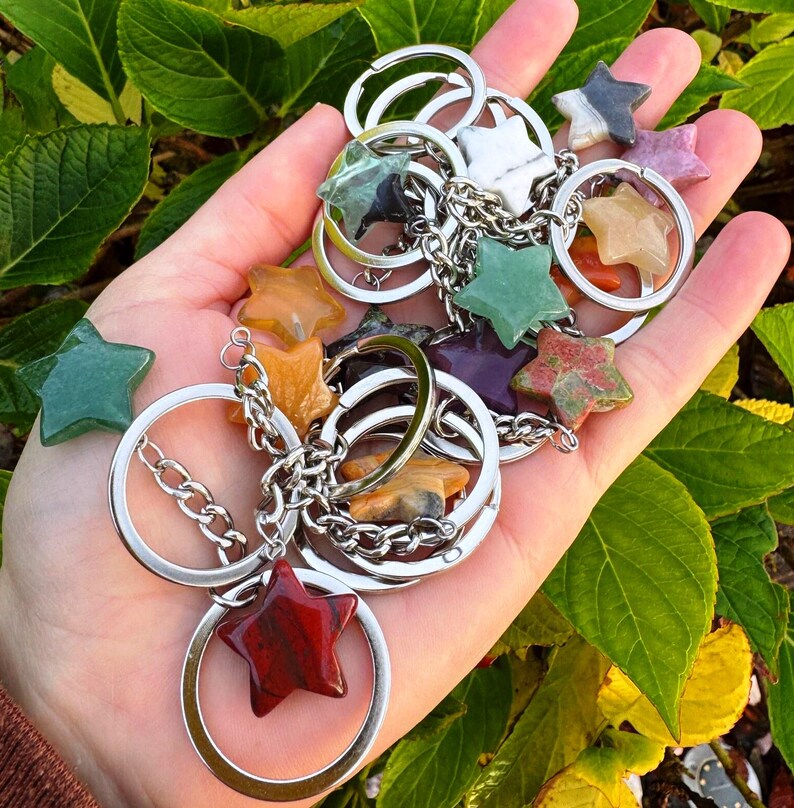 May include: A selection of star-shaped gemstone keyrings in various colours, including green, orange, red, and purple. Each star is attached to a silver keyring with a chain. The keyrings are held in a hand against a backdrop of green leaves.