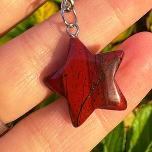 May include: A red star-shaped pendant with a silver chain. The pendant is made of a polished, reddish-brown stone with dark veins. The star is held up by a silver chain.