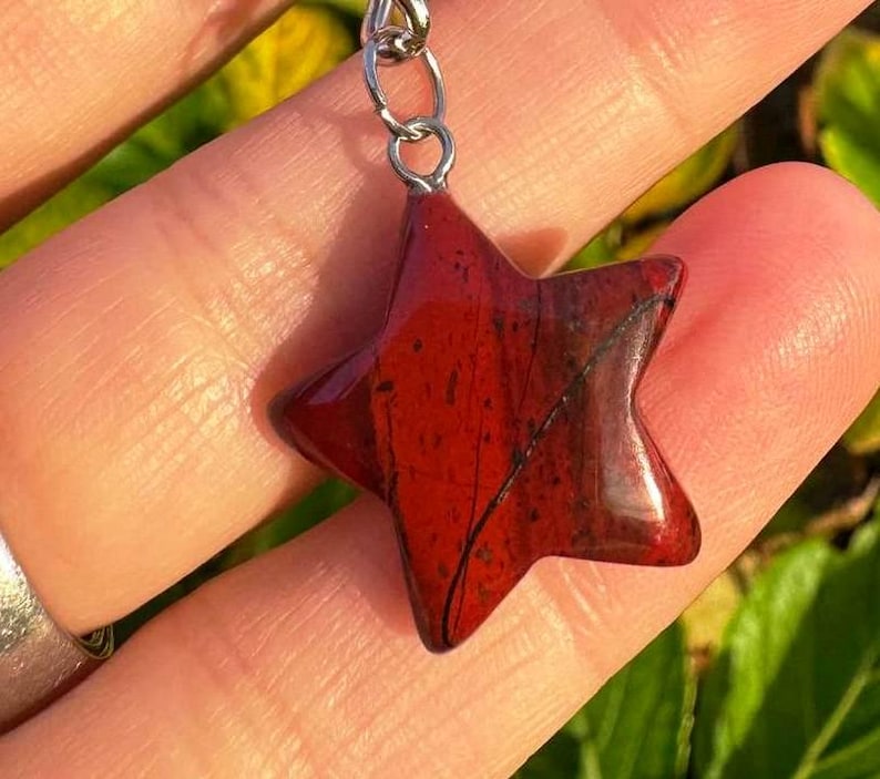 May include: A red star-shaped pendant with a silver chain. The pendant is made of a polished, reddish-brown stone with dark veins. The star is held up by a silver chain.