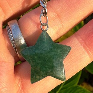 May include: A green star-shaped pendant made of polished stone, suspended from a silver-coloured chain. The pendant is held in a hand, with a silver ring with small clear stones on a finger. The background is blurred green foliage.