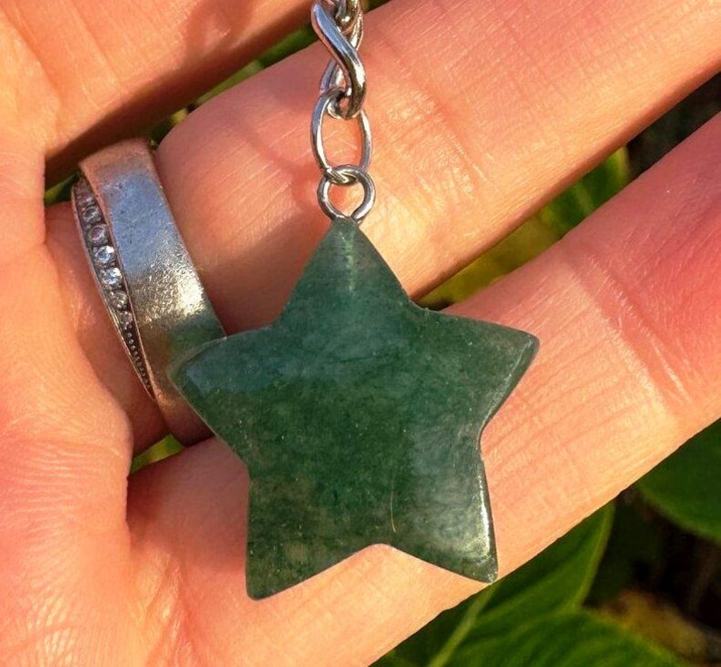 May include: A green star-shaped pendant made of polished stone, suspended from a silver-coloured chain. The pendant is held in a hand, with a silver ring with small clear stones on a finger. The background is blurred green foliage.