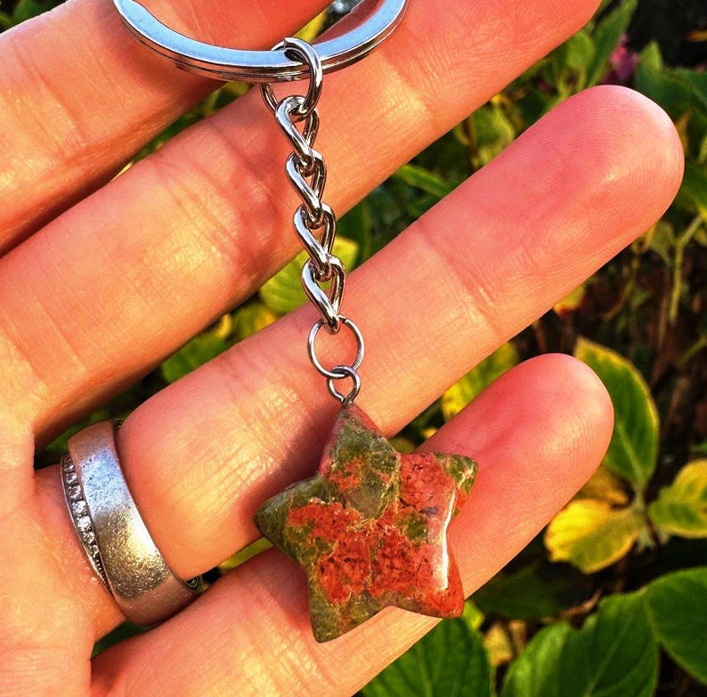 May include: A star-shaped charm made from red and green stone, attached to a silver chain and keyring. The background is blurred, showing green and yellow foliage. The charm is a keychain accessory.