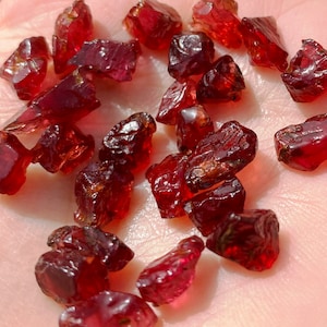 May include: A collection of small, rough, red gemstones. The gemstones are various shapes and sizes, and have a natural, unpolished look.