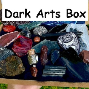 May include: A box filled with various dark arts themed items, including crystals, a small bottle, a black pentagram charm, and a red and black obsidian shard. The box is labeled "Dark Arts Box".