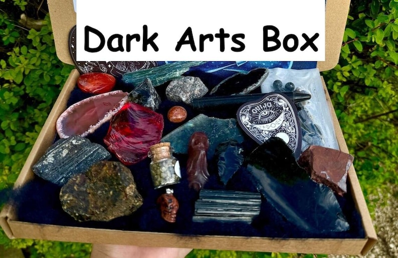 May include: A box filled with various dark arts themed items, including crystals, a small bottle, a black pentagram charm, and a red and black obsidian shard. The box is labeled "Dark Arts Box".