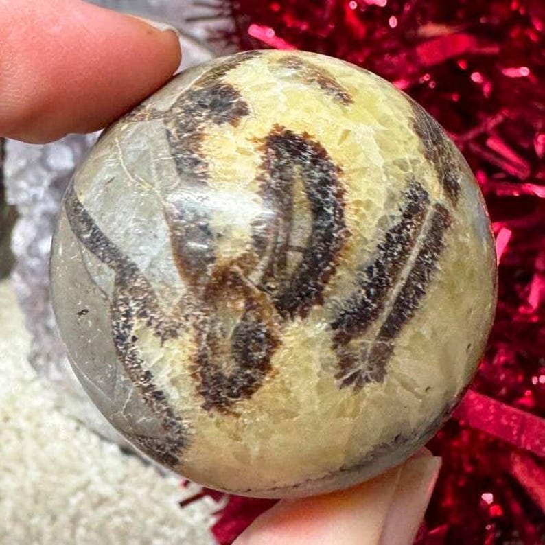 May include: A spherical decorative stone with a mix of tan, brown, and gray patterns. The polished surface of the stone is smooth and reflective. The stone is held by a person's fingers.