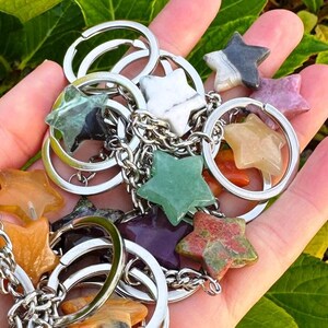 May include: A selection of star-shaped gemstone keyrings in diverse colours, such as green, orange, purple, and white. Each star is connected to a silver keyring with a chain. The keyrings are held in a hand against a background of green foliage.
