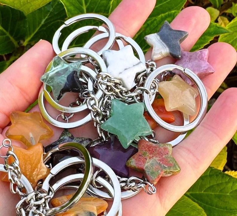 May include: A selection of star-shaped gemstone keyrings in diverse colours, such as green, orange, purple, and white. Each star is connected to a silver keyring with a chain. The keyrings are held in a hand against a background of green foliage.