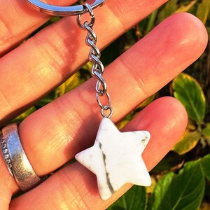 May include: A white star-shaped keychain charm with grey veining, attached to a silver chain and keyring. The charm is held in a hand, with a silver ring visible. The background is blurred with green and yellow foliage.
