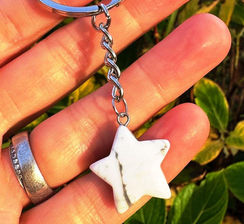 May include: A white star-shaped keychain charm with grey veining, attached to a silver chain and keyring. The charm is held in a hand, with a silver ring visible. The background is blurred with green and yellow foliage.