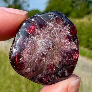 Peut inclure: Une pierre de grenat polie, rouge foncé, avec un intérieur cristallin translucide. La pierre a une forme lisse et arrondie. La pierre précieuse reflète la lumière, mettant en valeur sa couleur profonde et sa texture naturelle.