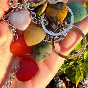 May include: A collection of heart-shaped gemstone keychains in various colors, including pink, red, yellow, green, and brown. Each heart is attached to a silver chain and keyring. The keychains are held in a hand, with green foliage in the background.