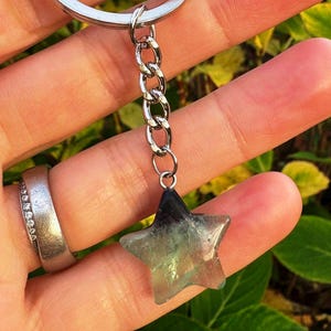 May include: A star-shaped keyring charm with a silver chain. The charm is made of a translucent, multi-coloured stone, with shades of green, blue, and black. The keyring is held by a person.