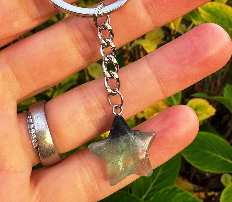 May include: A star-shaped keyring charm with a silver chain. The charm is made of a translucent, multi-coloured stone, with shades of green, blue, and black. The keyring is held by a person.