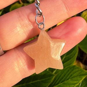 May include: A star-shaped pendant in a light orange hue, suspended from a silver-coloured chain. The pendant is smooth and polished, with a subtle shimmer. The background features green foliage.