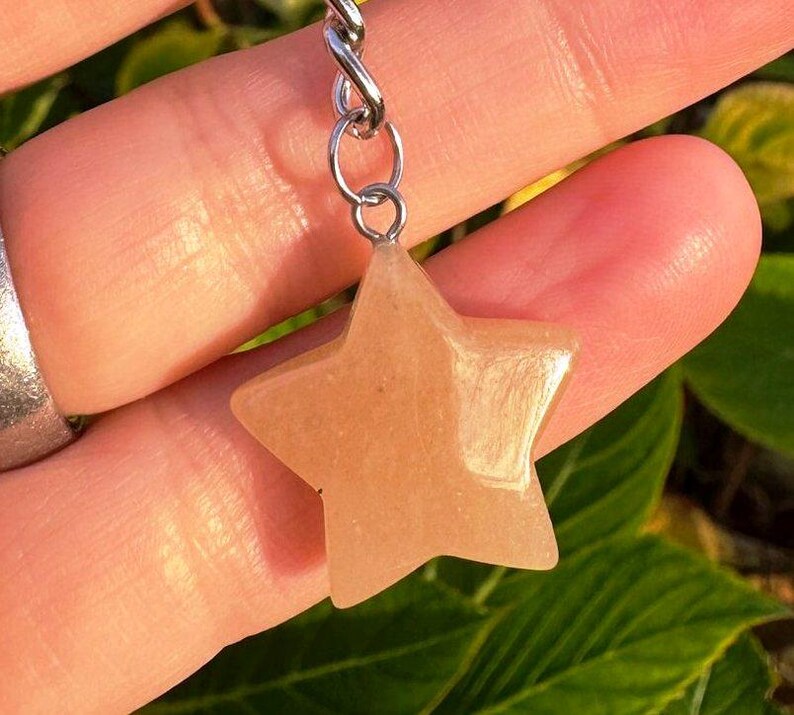 May include: A star-shaped pendant in a light orange hue, suspended from a silver-coloured chain. The pendant is smooth and polished, with a subtle shimmer. The background features green foliage.