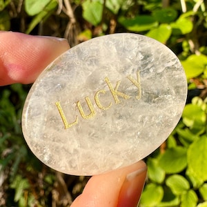 Peut inclure: Une pierre de cristal translucide de forme ovale avec le mot "Lucky" en lettres dorées. La pierre est tenue entre deux doigts, avec un arrière-plan vert flou de feuillage.