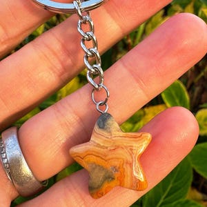 Op de afbeelding: Een stervormige sleutelhanger hanger van steen met oranje, bruine en grijze patronen. De hanger is bevestigd aan een zilveren ketting en sleutelring. De steen heeft een gepolijste afwerking en een natuurlijk uiterlijk.