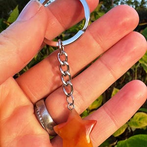 May include: A star-shaped keyring with a silver chain and ring. The star is a mix of orange and yellow hues. The keyring is held up against a background of green foliage.