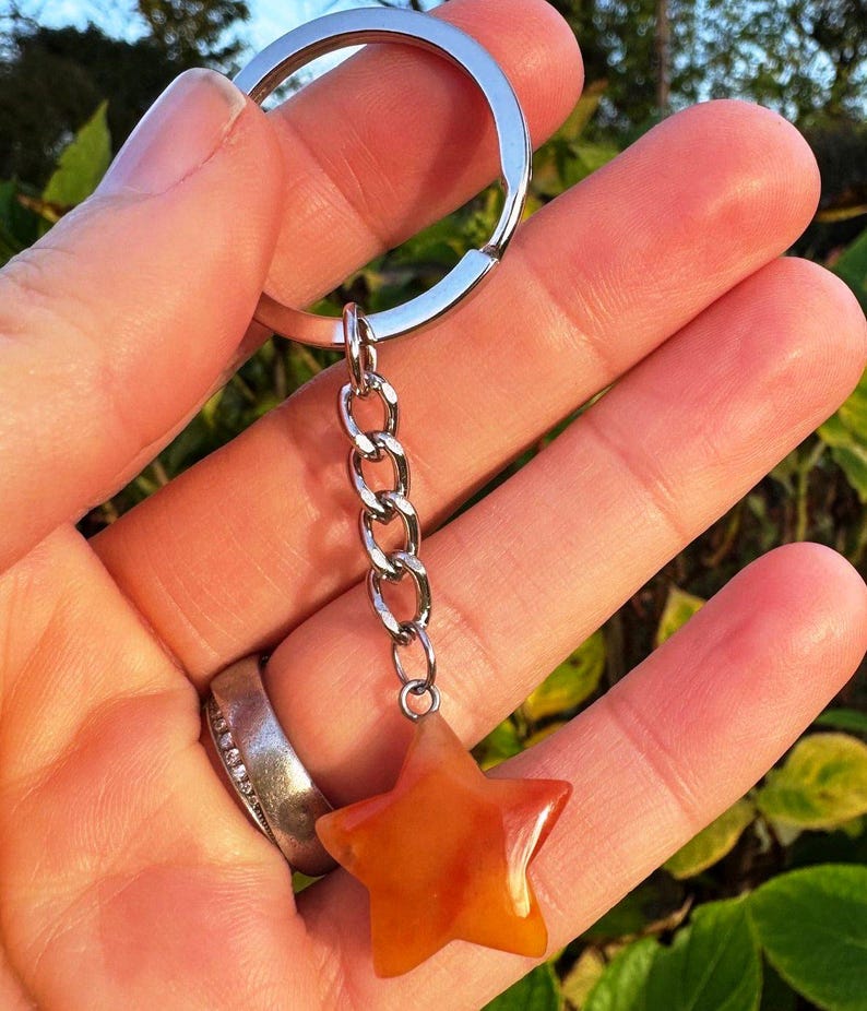 May include: A star-shaped keyring with a silver chain and ring. The star is a mix of orange and yellow hues. The keyring is held up against a background of green foliage.
