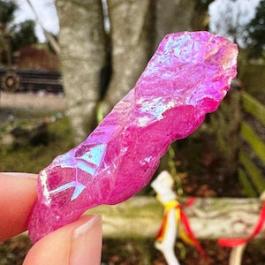 May include: A striking, pink crystal with shimmering, rainbow-like colours. The raw gemstone is held in a hand, highlighting its natural, textured surface. The crystal's colour varies from deep magenta to lighter pink shades, with areas of blue and green iridescence.