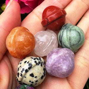 May include: A collection of polished gemstones in various colors and patterns. The assortment includes orange, clear, green, purple, and speckled stones, each with a unique shape and texture. The gemstones are held in a hand, showcasing their natural beauty.