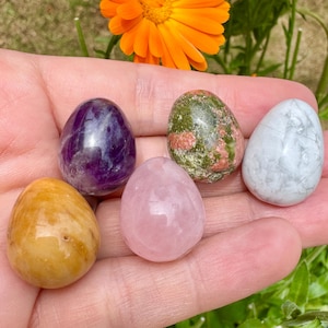 Puede incluir: Colección de seis huevos de piedras preciosas pulidas en varios colores, incluyendo púrpura, rosa, amarillo, verde y blanco. Los huevos se sostienen en una mano, con una flor naranja brillante en el fondo. Las piedras tienen un acabado liso y brillante.