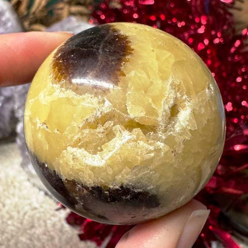 May include: A polished sphere with a mix of yellow, brown, and black hues. The stone has a textured surface and a rounded shape. The sphere is held in a person's hand.