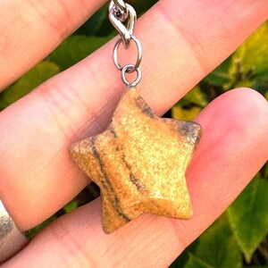 May include: A star-shaped pendant made of tan and brown stone, suspended from a silver chain. The stone has natural patterns and textures. The pendant is a small, decorative accessory.