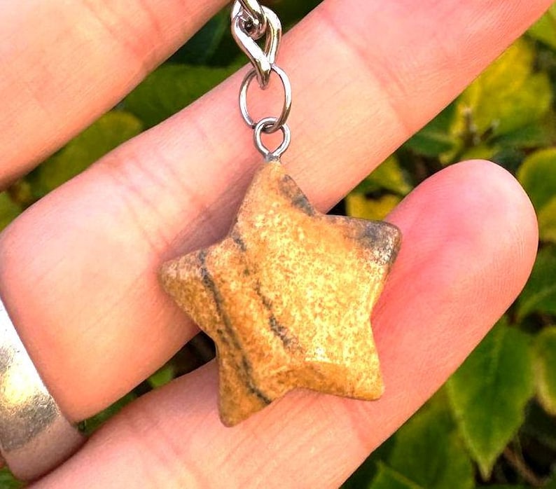 May include: A star-shaped pendant made of tan and brown stone, suspended from a silver chain. The stone has natural patterns and textures. The pendant is a small, decorative accessory.