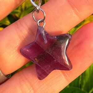May include: A star-shaped pendant crafted from purple fluorite, hanging from a silver-coloured chain. The gemstone is translucent, displaying internal patterns. The pendant is held against a backdrop of skin and foliage.
