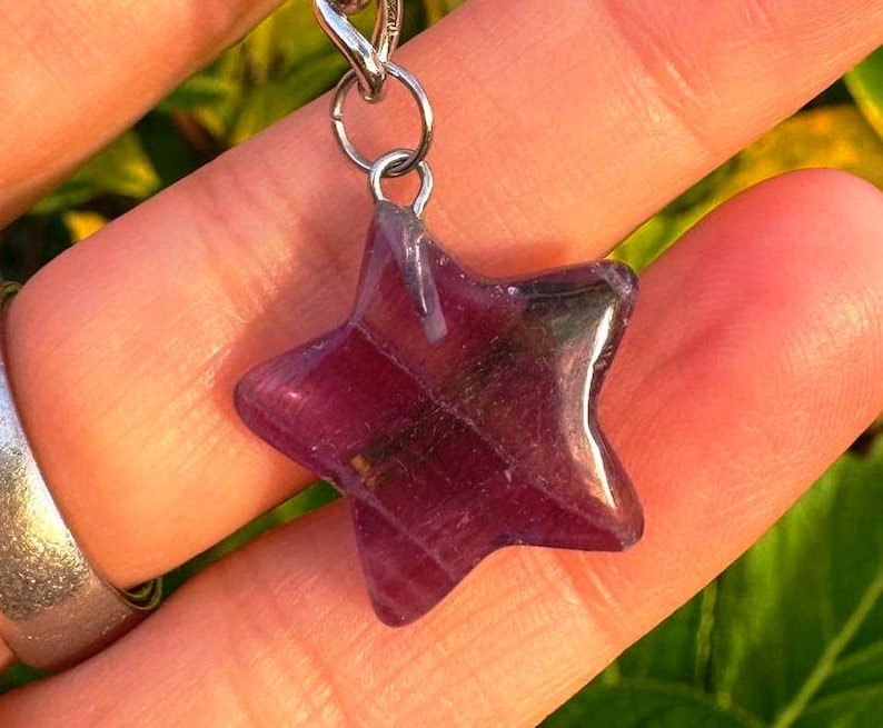 May include: A star-shaped pendant crafted from purple fluorite, hanging from a silver-coloured chain. The gemstone is translucent, displaying internal patterns. The pendant is held against a backdrop of skin and foliage.
