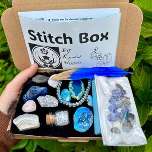 May include: A Stitch Box, open to reveal a collection of crystals, a blue feather, and a beaded necklace. The box contains a variety of blue and gray stones, a small glass bottle, and a bag of small crystals. The box has the text "Stitch Box" and "Elf Kendal Hippies".