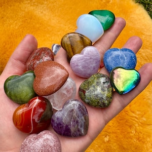 以下が含まれることがあります： さまざまな色のハート型の宝石のコレクションが手に持たれています。緑、赤、紫、青、金などがあります。宝石はさまざまな質感と模様を示し、一部は半透明で、一部は不透明です。背景はぼやけたオレンジ色の表面です。