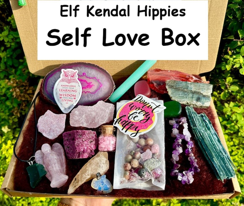 May include: A self-love box filled with crystals, a pink agate slice, a small bottle of pink crystals, a green stone pendant, a pink angel figurine, a sticker that says "Don't worry be happy", and a purple amethyst necklace. The box is labeled "Elf Kendal Hippies Self Love Box".