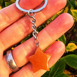 May include: A star-shaped keyring with a silver chain and ring. The star is a warm orange colour and appears to be made of a smooth, polished material. The background is blurred, with green and yellow foliage.