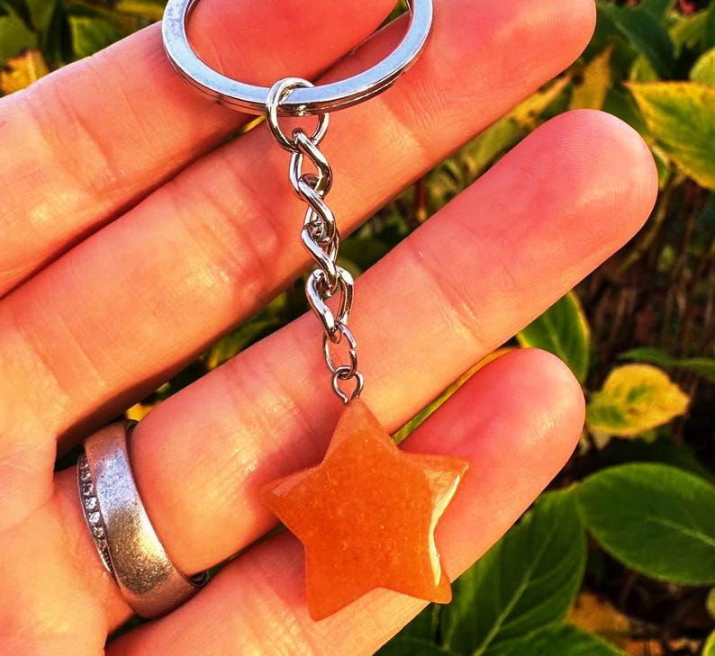 May include: A star-shaped keyring with a silver chain and ring. The star is a warm orange colour and appears to be made of a smooth, polished material. The background is blurred, with green and yellow foliage.