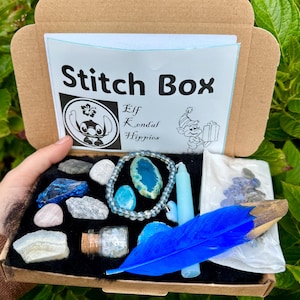 May include: A brown cardboard box labeled "Stitch Box" with various blue and white crystals, a blue feather, a blue candle, and a bracelet. The box also contains a small glass bottle and a small bag of crystals. The box is open, revealing its contents.
