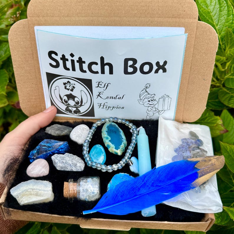May include: A brown cardboard box labeled "Stitch Box" with various blue and white crystals, a blue feather, a blue candle, and a bracelet. The box also contains a small glass bottle and a small bag of crystals. The box is open, revealing its contents.