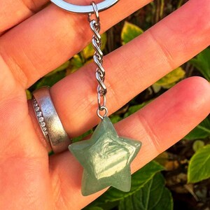 May include: A green star-shaped gemstone keyring. The star is suspended from a silver-coloured chain and ring. The gemstone has a translucent quality, and the star's five points are clearly defined. The background is blurred, suggesting an outdoor setting.