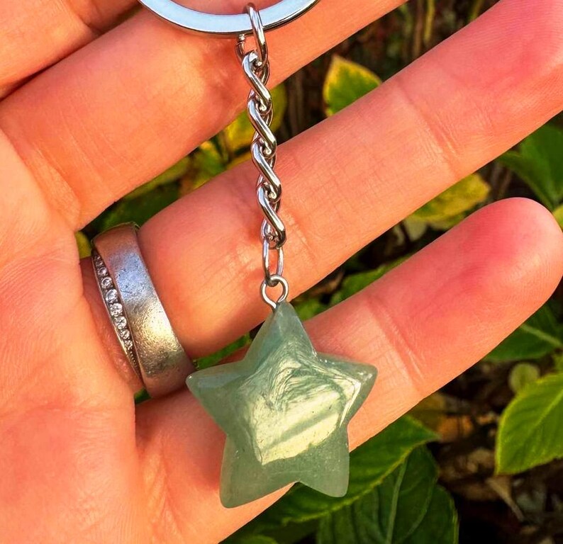 May include: A green star-shaped gemstone keyring. The star is suspended from a silver-coloured chain and ring. The gemstone has a translucent quality, and the star's five points are clearly defined. The background is blurred, suggesting an outdoor setting.