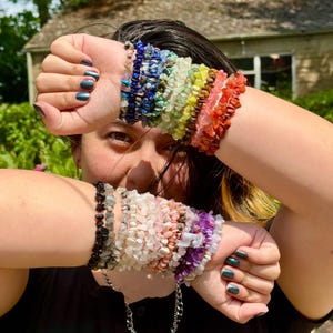 Puede incluir: Una colección de pulseras de virutas de piedras preciosas en varios colores, incluyendo azul, verde, amarillo, naranja y rojo. Las pulseras están apiladas en las muñecas, mostrando un arcoíris de joyería de piedra natural. Las pulseras están hechas de piedras pequeñas de forma irregular.