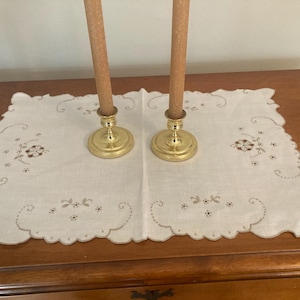 May include: Two brass candle holders with brown candles on a white embroidered table runner.
