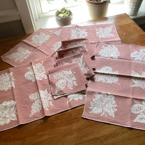 May include: A collection of pink linen napkins and placemats with white floral designs. The napkins and placemats are arranged on a wooden table, showcasing various botanical patterns. The set includes different sizes and shapes, perfect for a dining setting.
