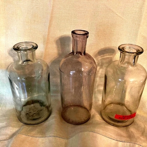 May include: Three vintage glass bottles in different shapes and sizes. The bottles are clear with grey and purple tints. One bottle has a red label near the base. They are displayed on a cream-coloured fabric.