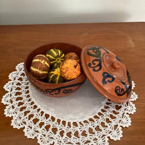 Könnte beinhalten: Eine terrakottafarbene Keramikschale mit Deckel, verziert mit blauen und schwarzen Wirbeln, steht auf einer weißen Spitzendecke. Die Schale enthält mehrere kleine, bunte Kürbisse in Orange-, Gelb- und Grüntönen.