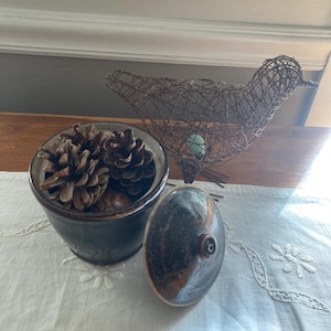May include: A decorative arrangement featuring a dark blue ceramic pot filled with pine cones, a wire bird sculpture, and a ceramic lid. The bird sculpture has a teal-coloured egg inside. The items are arranged on a white embroidered runner.