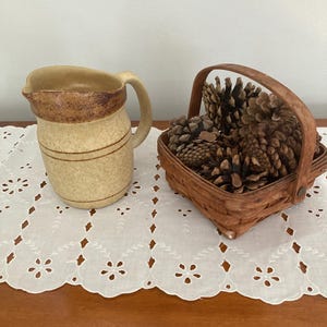 Könnte beinhalten: Eine beige Keramikkrug mit Henkel und braunen Akzenten steht auf einer weißen Lochspitzentischdecke. Ein kleiner, geflochtener Korb mit Tannenzapfen befindet sich neben dem Krug. Die Gegenstände stehen auf einer Holzoberfläche.