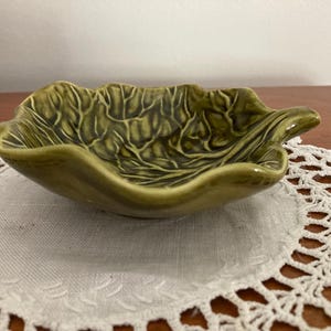 May include: A green ceramic leaf-shaped dish with detailed veining. The dish is a muted olive green color and sits on a white lace doily. The dish is likely for serving or display.